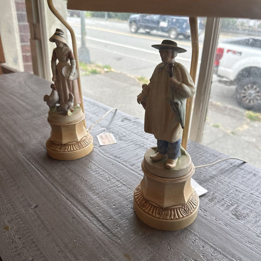 Two decorative lamps with figurines on a table, with a window view of a street.