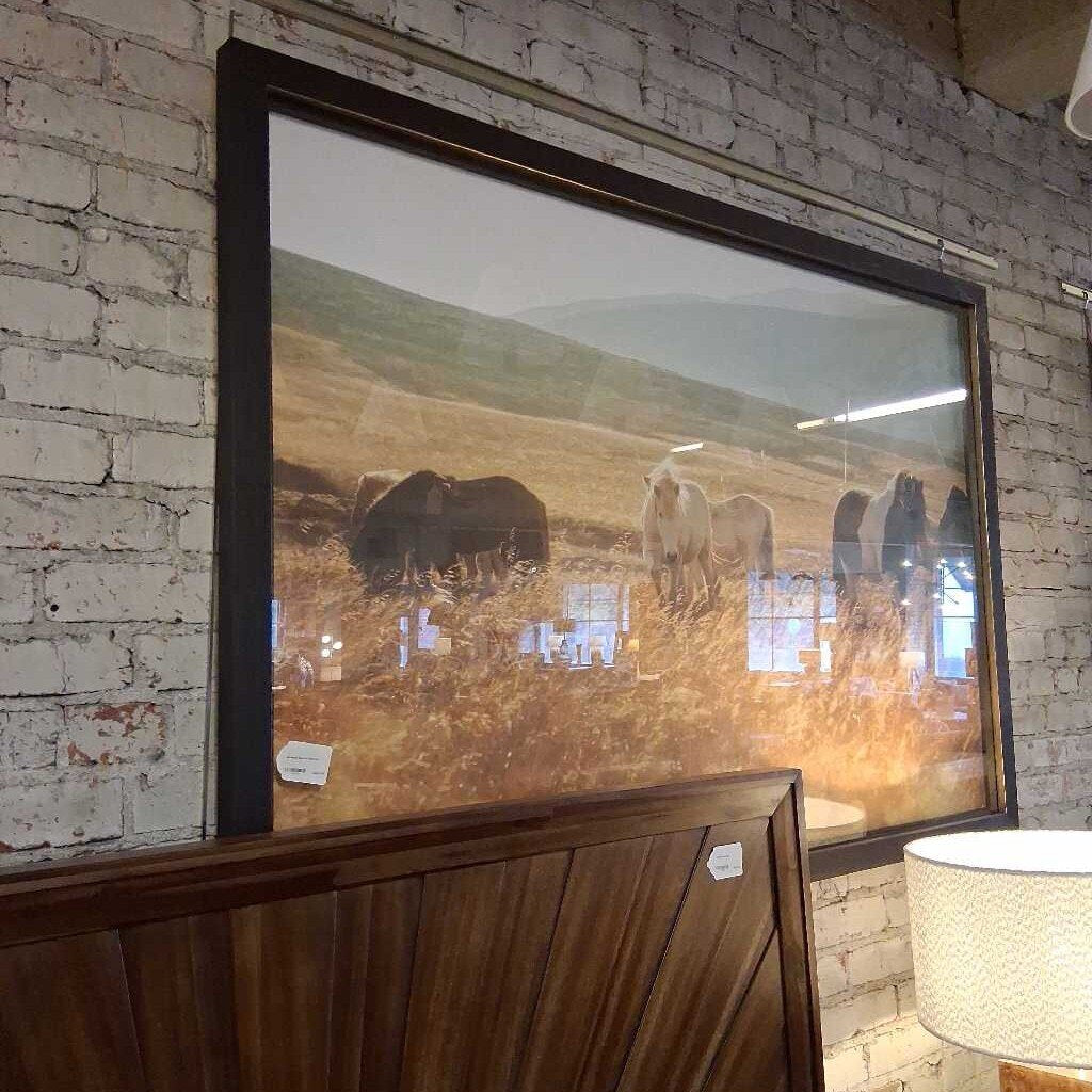 Framed painting of a landscape with animals on a brick wall.