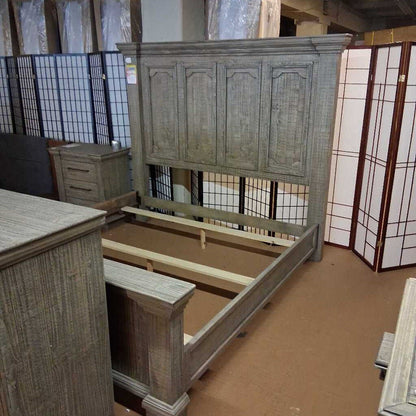 Wooden bed frame in a storage or warehouse setting with other furniture.