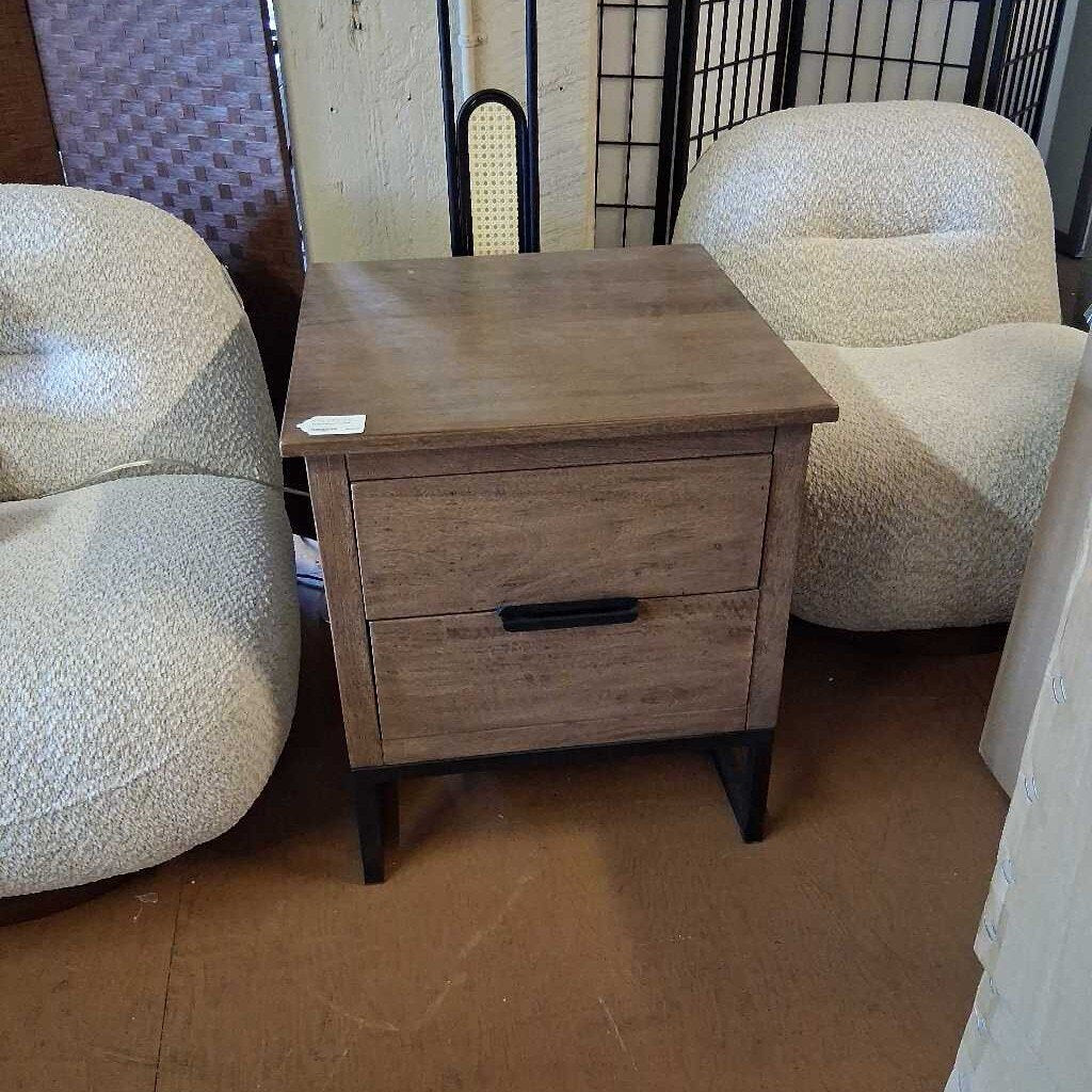 Wooden nightstand with two drawers in a room with patterned chairs.