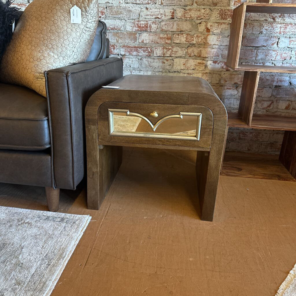 Wooden side table with decorative panel next to a brown leather chair against a brick wall.
