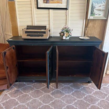 Dark wooden cabinet with open doors, featuring a typewriter and decorative items on top, in a room with a patterned rug and framed pictures on the wall.