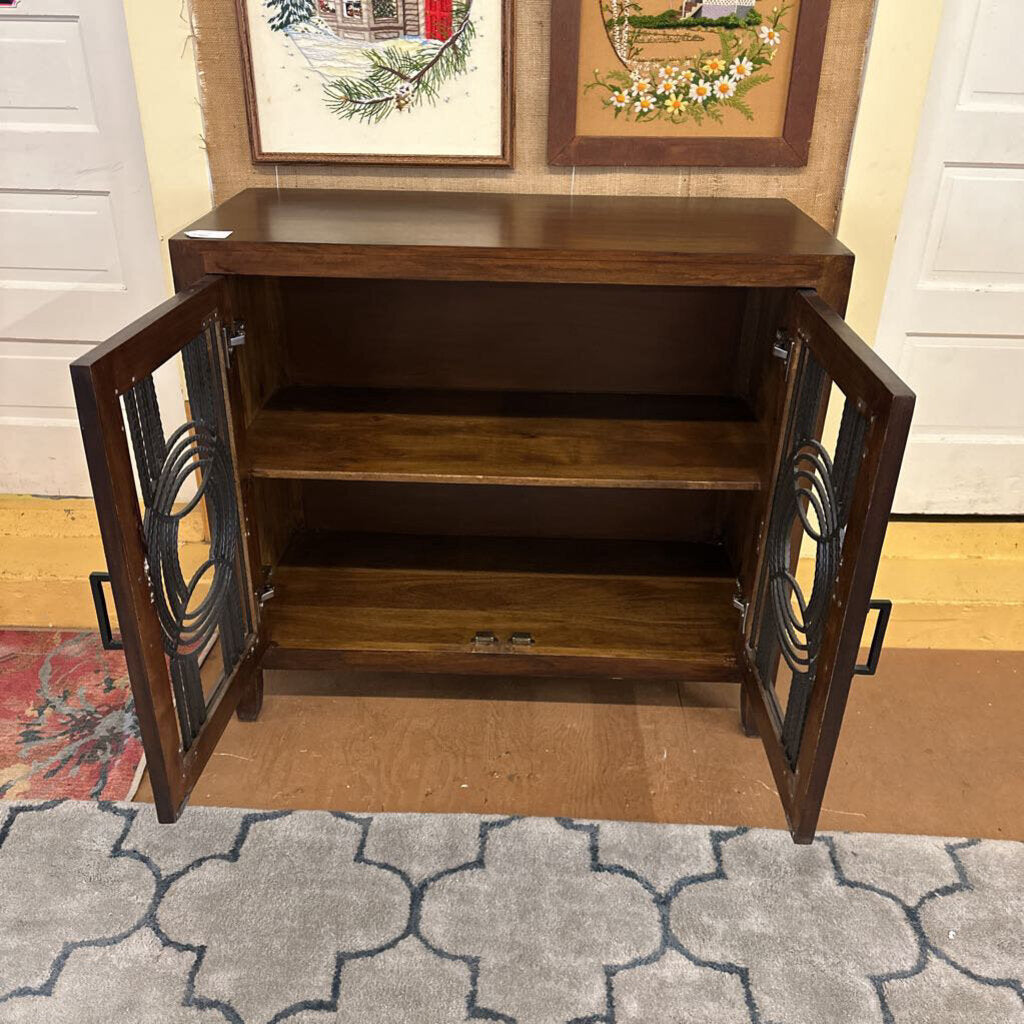 Wooden cabinet with glass doors on a carpeted floor, with two framed pictures on the wall in the background.