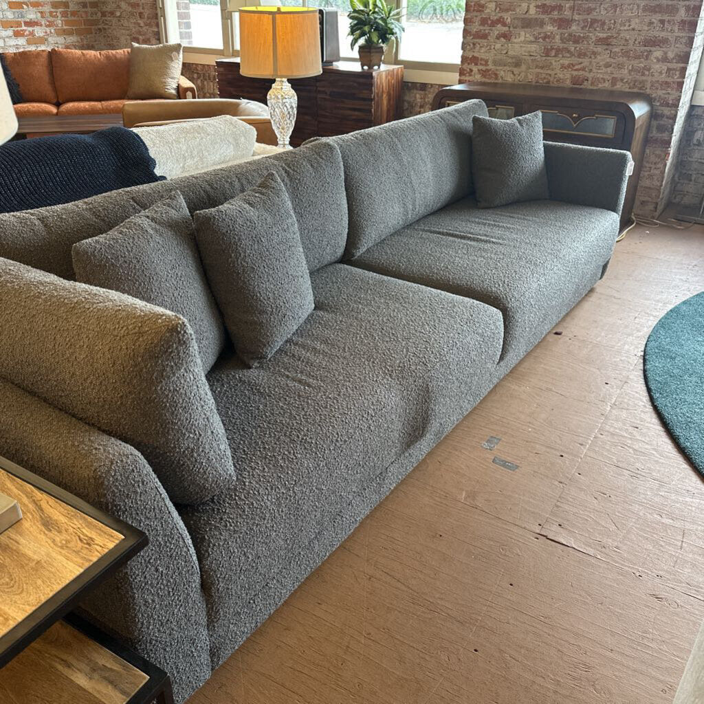 Gray sectional sofa in a living room setting with wooden floor and brick wall.