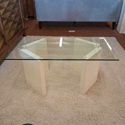 Glass coffee table on a beige carpet with a blue ottoman in the background
