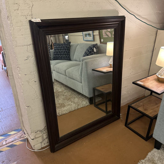 Large mirror with dark wood frame reflecting a living room with a sofa and lamp.