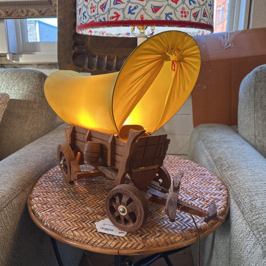 Wooden lamp shaped like a covered wagon with a yellow shade on a wicker table.