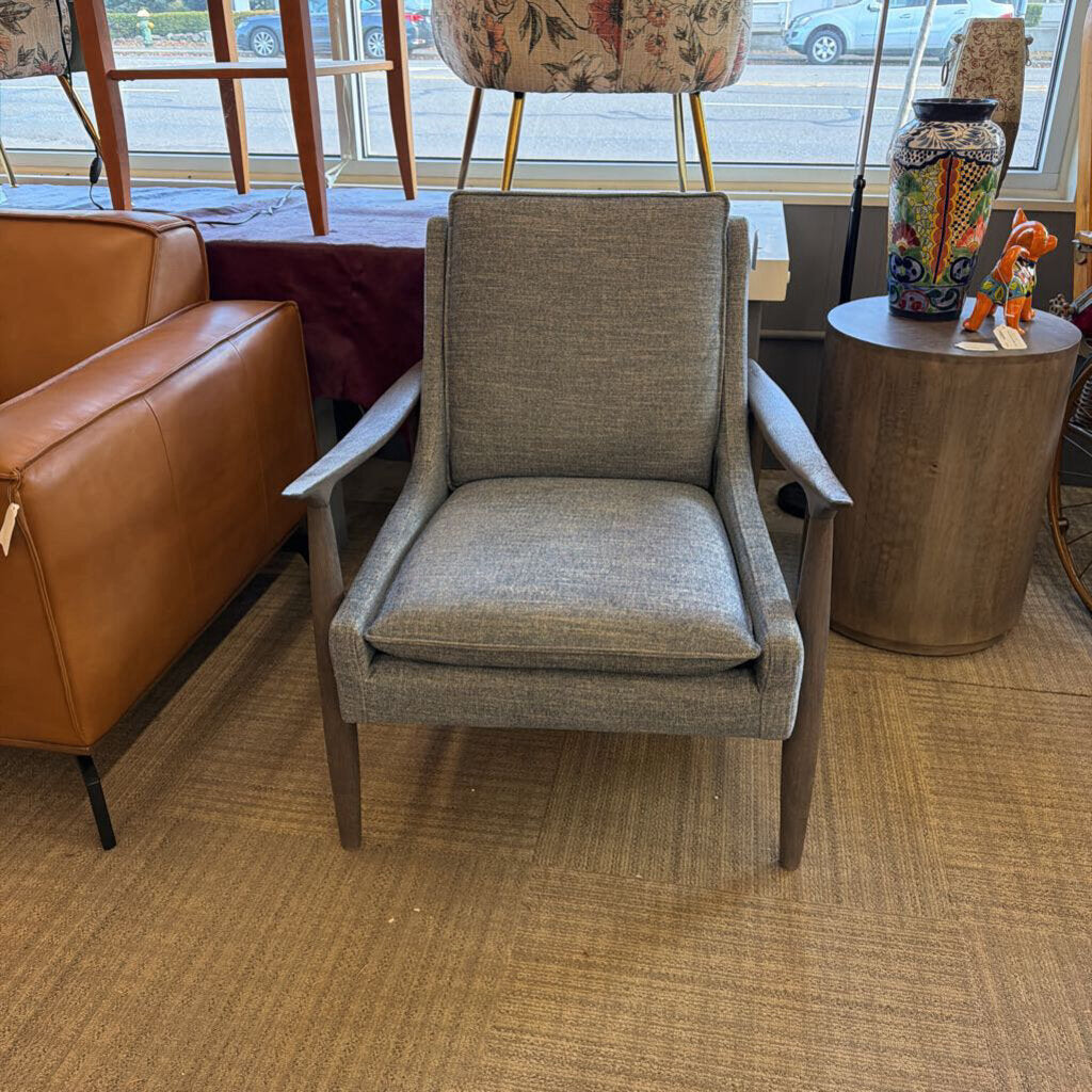 Gray upholstered chair in a room with a brown leather couch and a small round table.