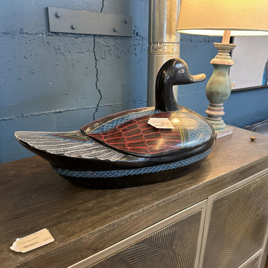 Decorative duck-shaped bowl on a wooden surface with a lamp in the background.