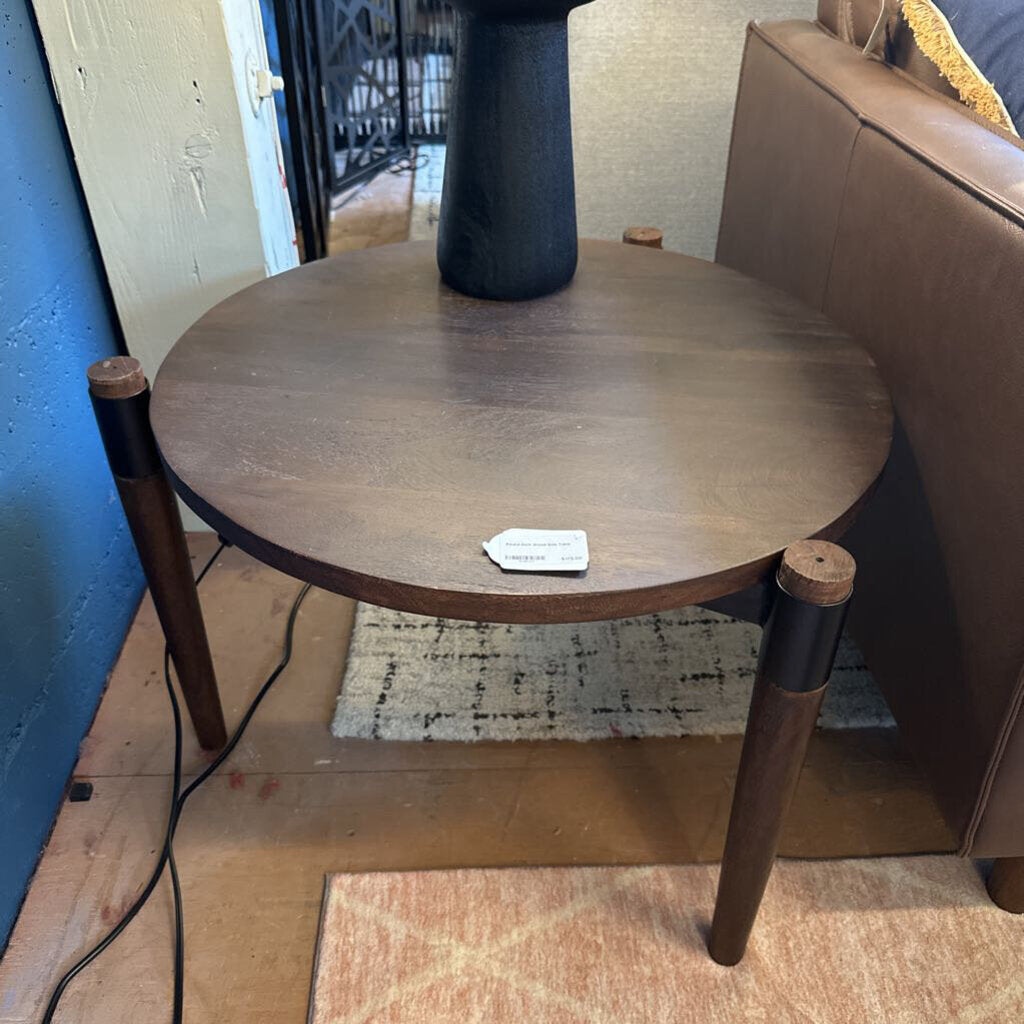 Round wooden side table with a tag on a wooden floor next to a brown sofa.