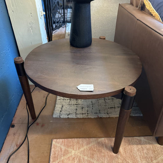 Round wooden side table with a tag on a wooden floor next to a brown sofa.