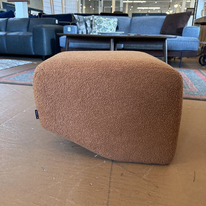 Brown textured ottoman on a wooden floor with a blurred background of furniture.