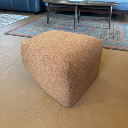 Brown textured cube-shaped ottoman on a wooden floor with a rug and table in the background.