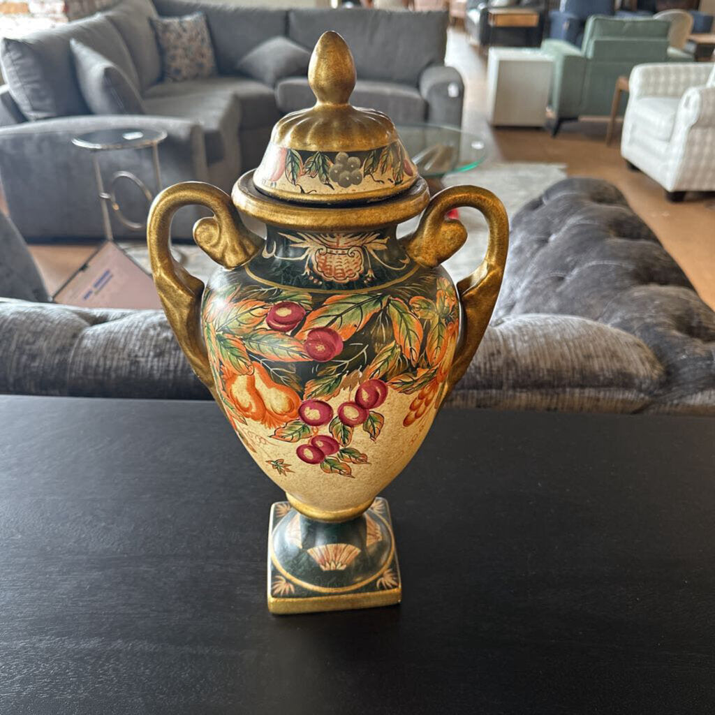 Decorative urn with floral and fruit design on a dark surface, with a living room in the background.