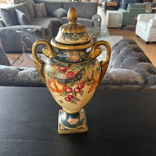 Decorative urn with floral and fruit design on a dark surface, with a living room in the background.