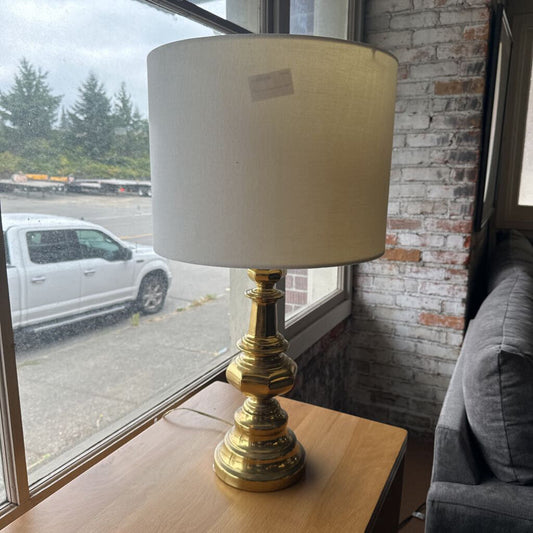 Lamp with a white shade and gold base on a wooden table near a window.
