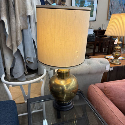 Brass table lamp with beige shade on a glass table in a room setting.