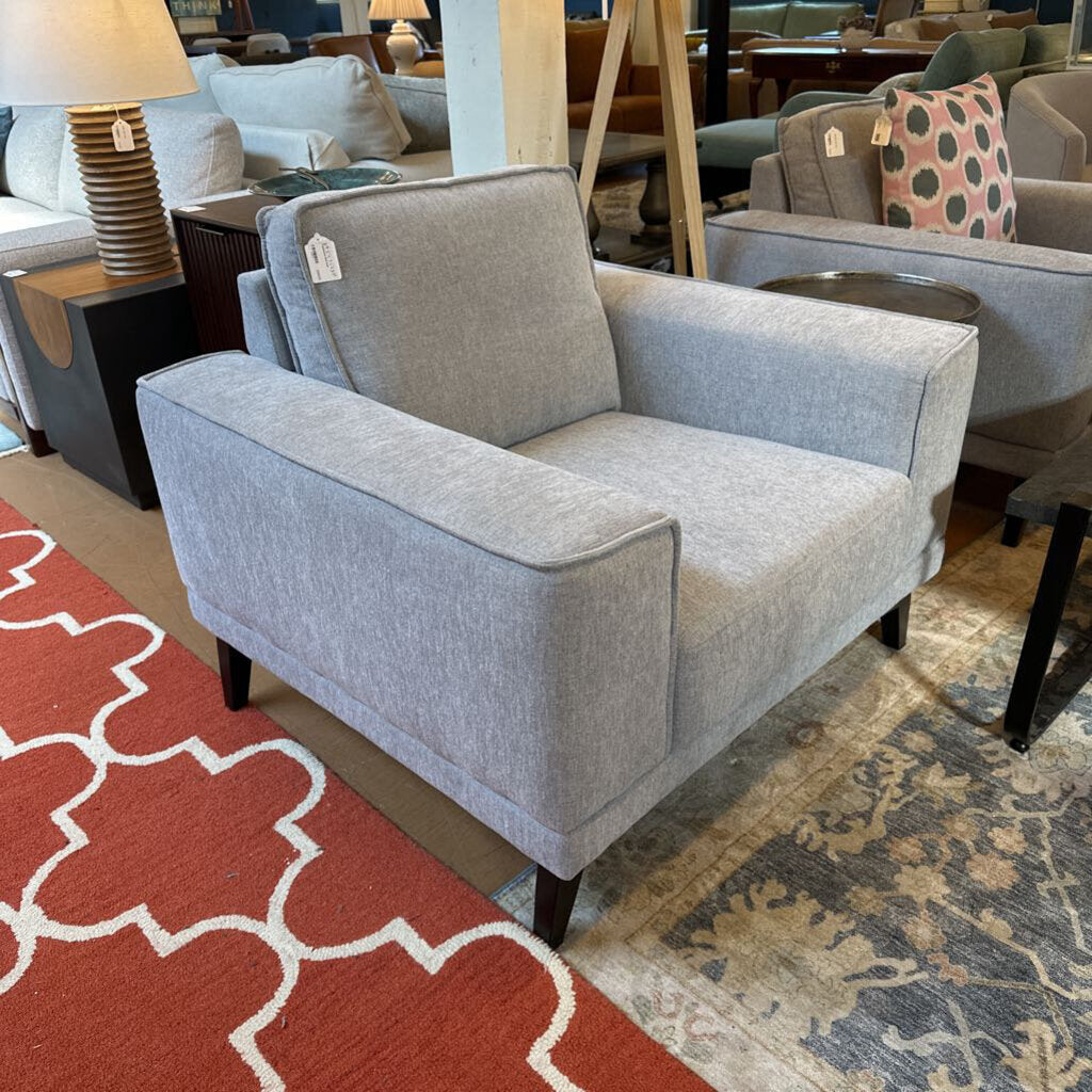 Gray armchair in a showroom with other furniture and a red patterned rug.