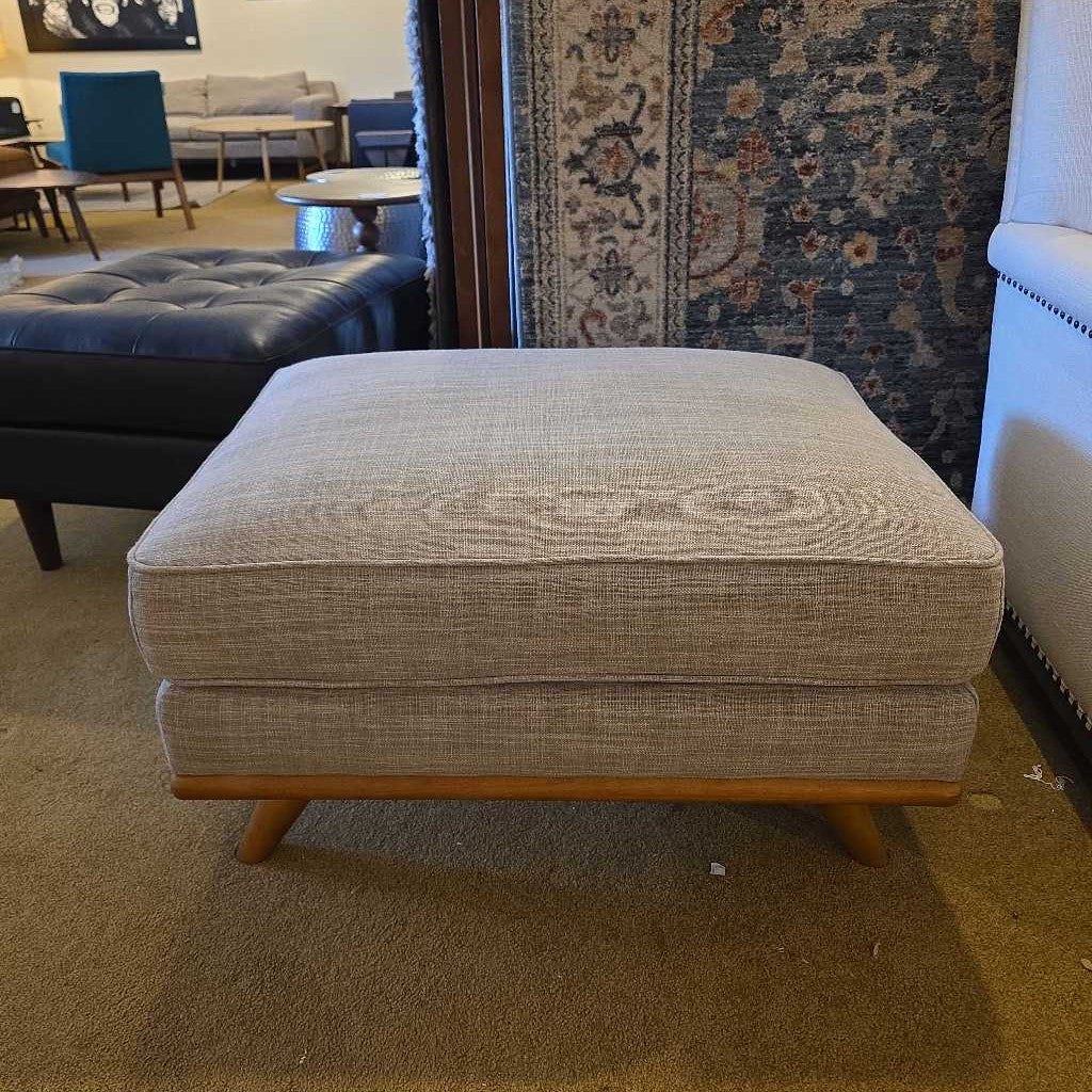 Beige ottoman with wooden legs in a room with furniture and a rug.