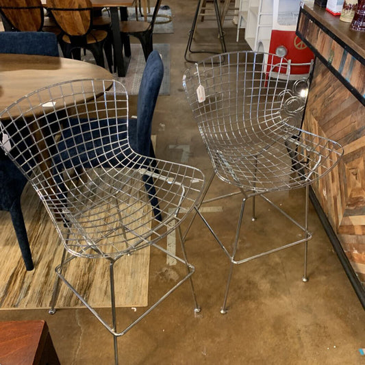 Two metal wire chairs on a wooden pallet in an indoor setting with a counter and stools in the background.
