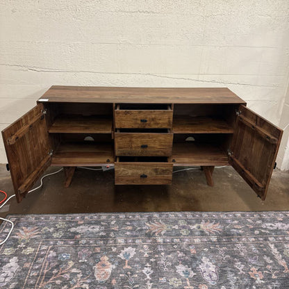 Wooden sideboard with open doors on a patterned rug against a white brick wall.