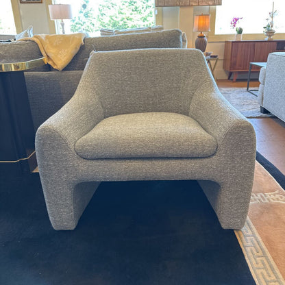 Gray armchair in a living room setting with a couch and lamps in the background.
