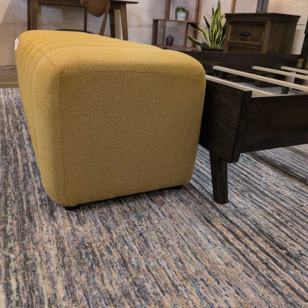 Yellow cube-shaped ottoman on a patterned carpet with a wooden chair and plant in the background.