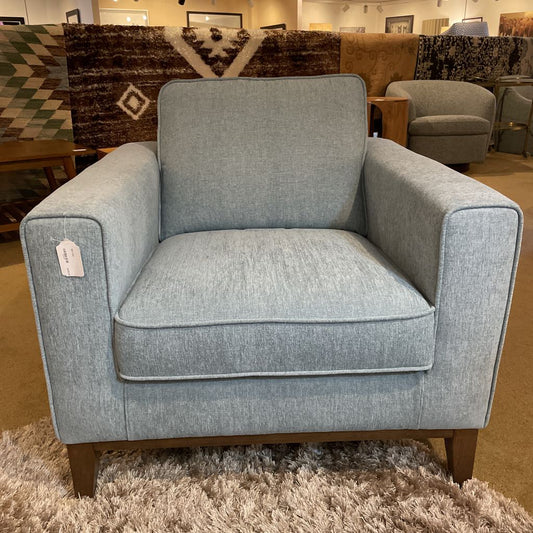 A light blue upholstered armchair with square arms, a single back cushion, and tapered wooden legs resting on a high-pile shag rug