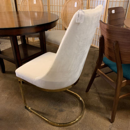White chair with gold frame on a concrete floor with other chairs and a table in the background.