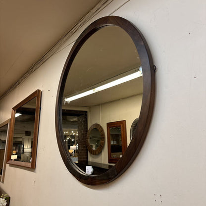 Round wooden-framed mirror on a wall with other mirrors in the background.