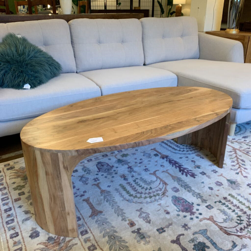 A light wood, oval-shaped coffee table with seamless curved waterfall edges, positioned on a decorative patterned rug in front of a light grey sectional sofa
