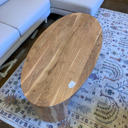 A light wood, oval-shaped coffee table with seamless curved waterfall edges, positioned on a decorative patterned rug in front of a light grey sectional sofa