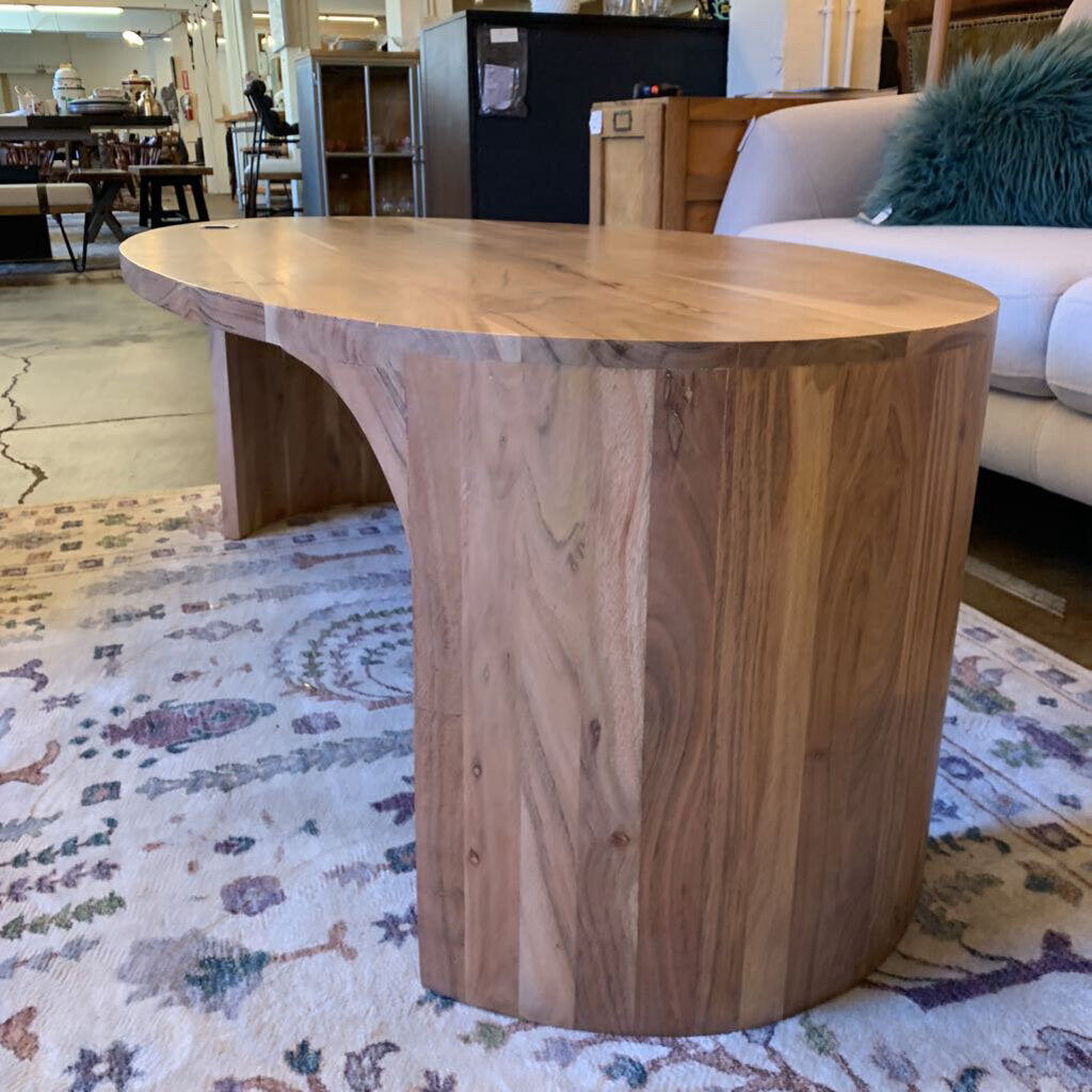 A light wood, oval-shaped coffee table with seamless curved waterfall edges, positioned on a decorative patterned rug in front of a light grey sectional sofa