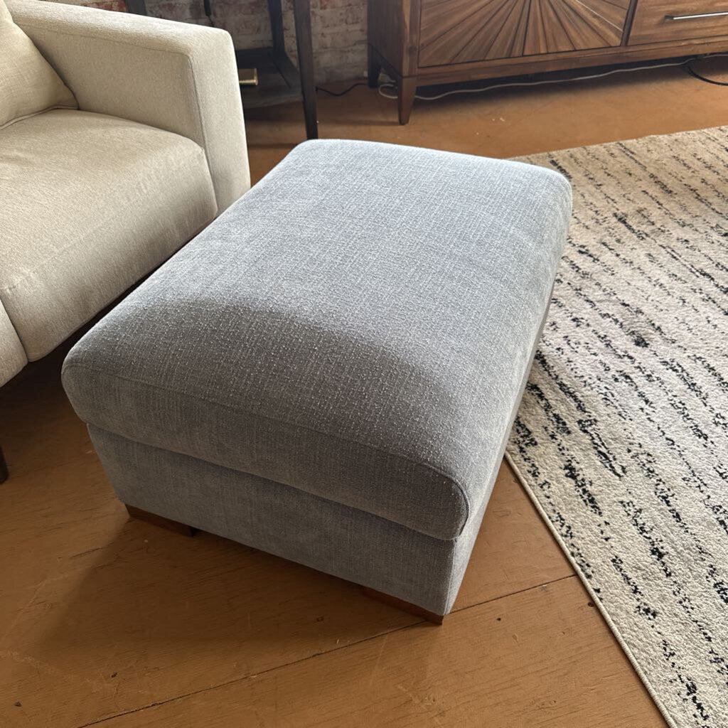 Gray ottoman in a living room setting with a beige sofa and wooden floor.