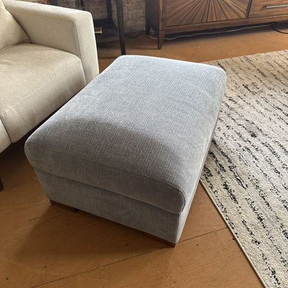 Gray ottoman in a living room setting with a beige sofa and wooden floor.