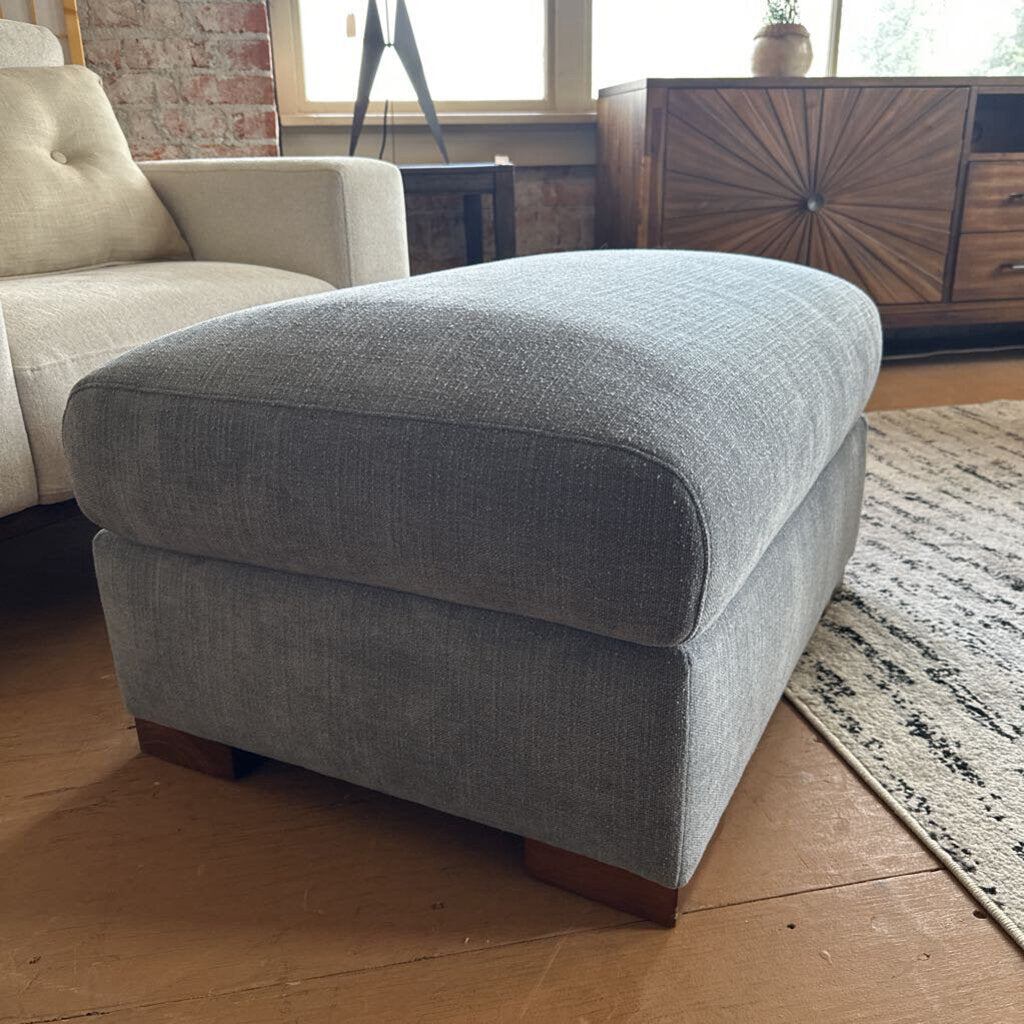 Gray upholstered ottoman in a living room setting with a beige armchair and wooden floor.