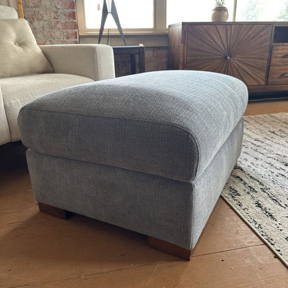Gray upholstered ottoman in a living room setting with a beige armchair and wooden floor.
