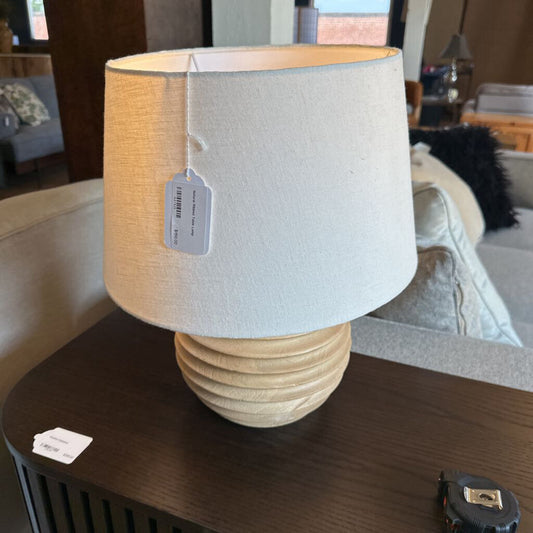 Lamp with a beige shade and textured base on a wooden surface, with a blurred indoor background.