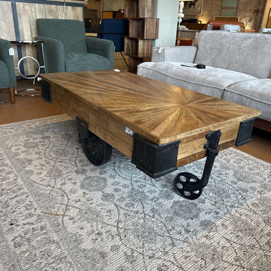 Wooden coffee table with starburst design and metal wheels in a furniture store setting.