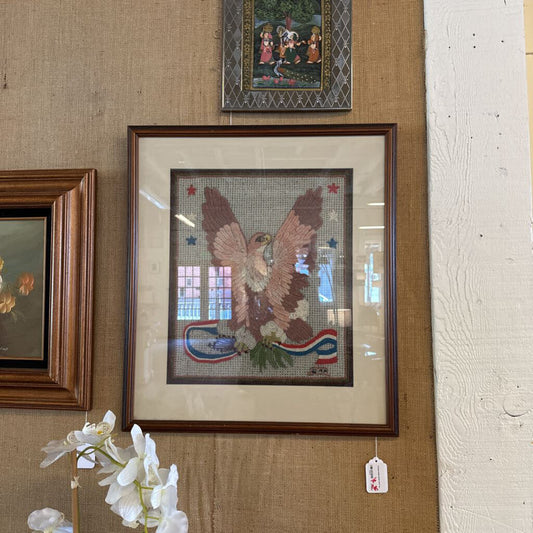 Framed cross-stitched artwork of an eagle on a textured wall with other framed pictures.