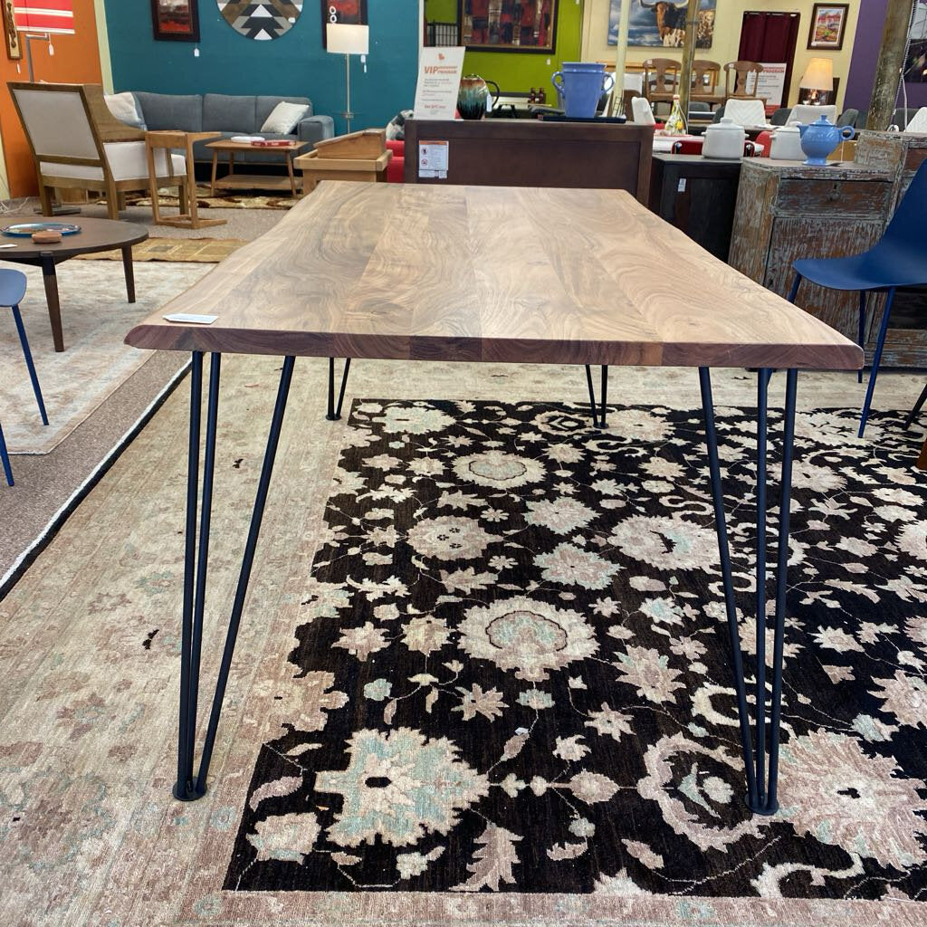 Wooden dining table with black hairpin legs on a patterned rug in a showroom setting.