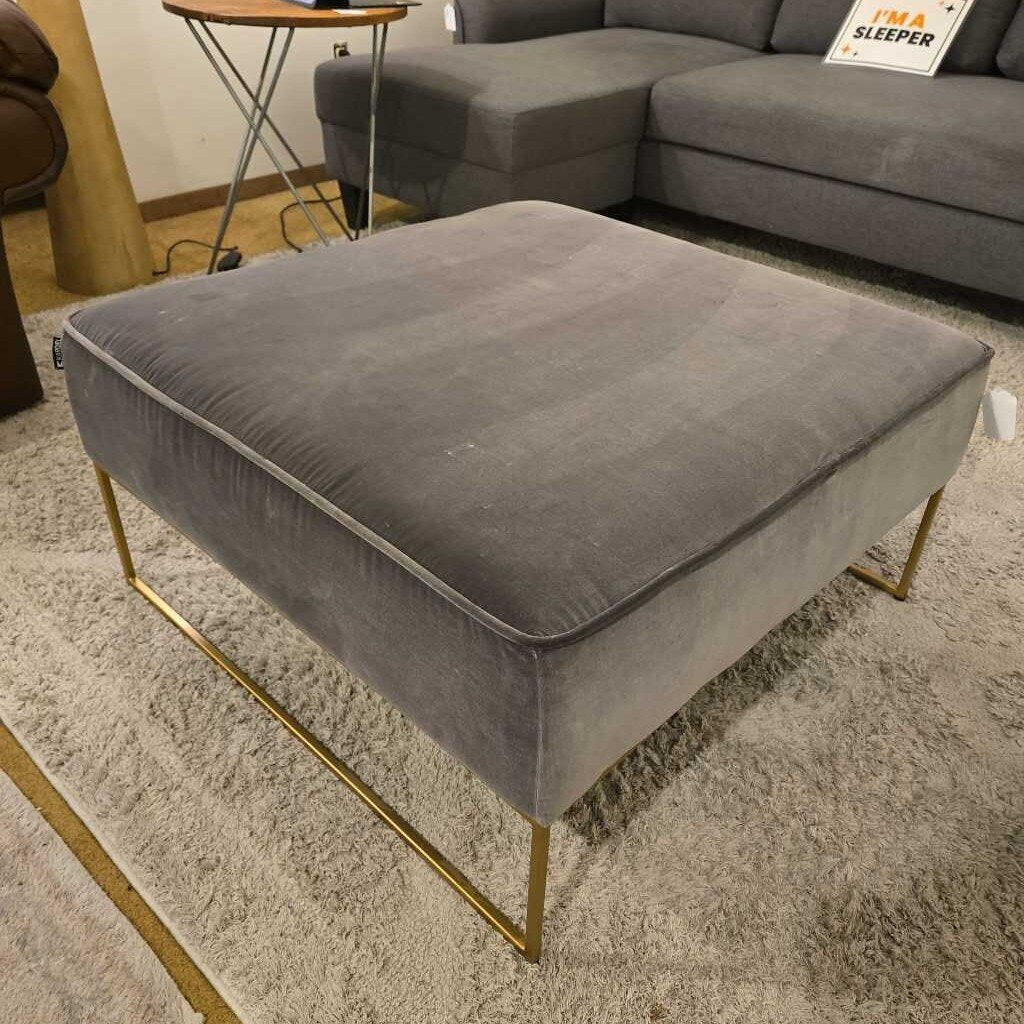 Gray ottoman with gold legs on a carpeted floor in a living room setting.