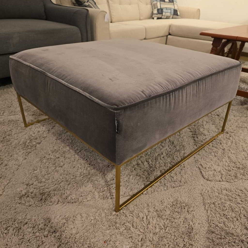 Gray ottoman with gold legs on a carpeted floor