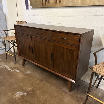 Wooden sideboard with drawers and doors in a room with chairs and a painting.