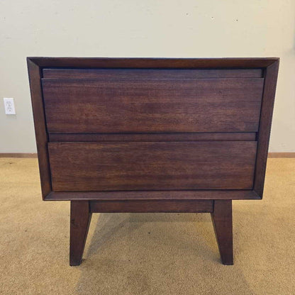 Wooden nightstand with two drawers on a beige carpeted floor.
