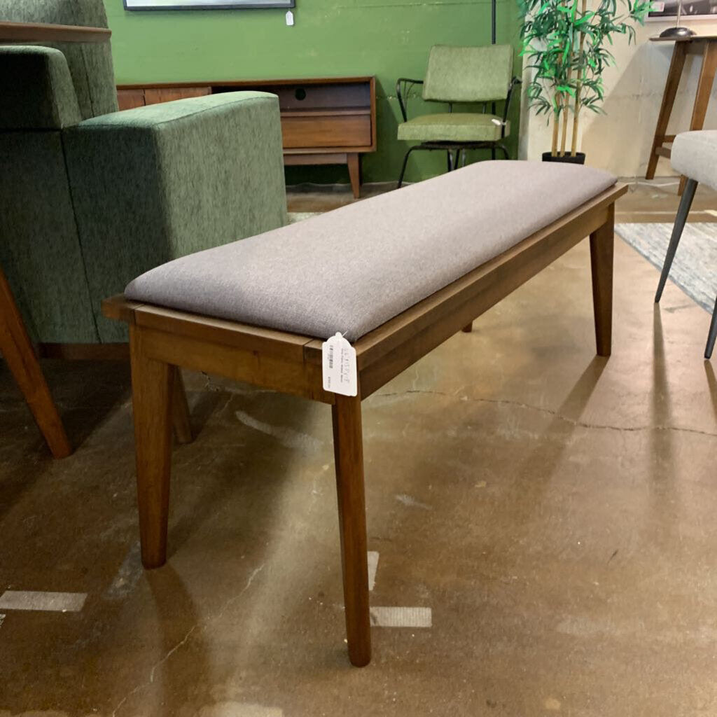 Wooden bench with a gray cushion on a tiled floor, surrounded by green chairs and a plant.