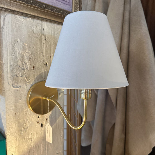 Wall-mounted lamp with a white shade and gold arm against a textured wall.