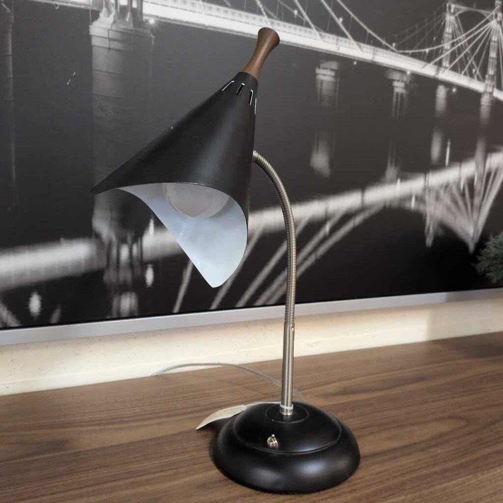 Black adjustable desk lamp on a wooden surface with a blurred background of a bridge.