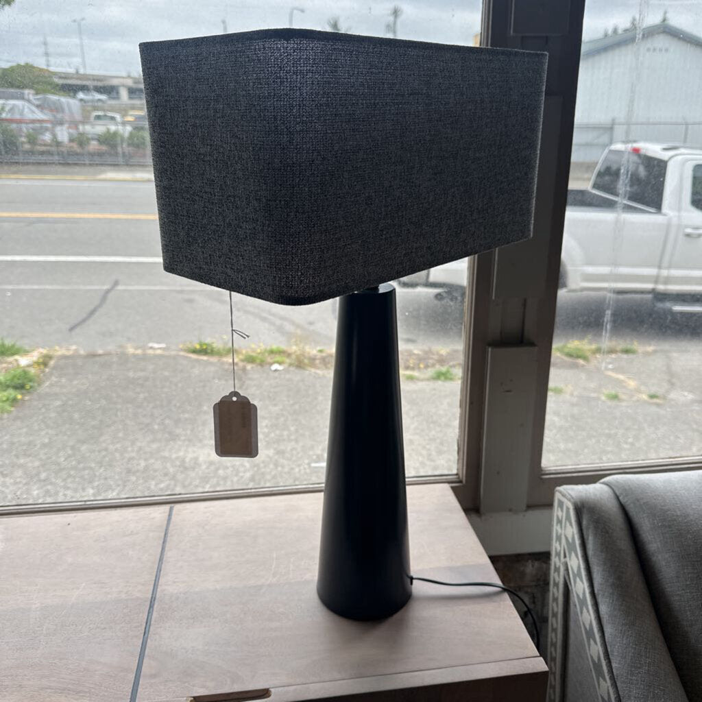 Black table lamp with a textured shade on a wooden surface, with a parking lot visible through a window in the background.