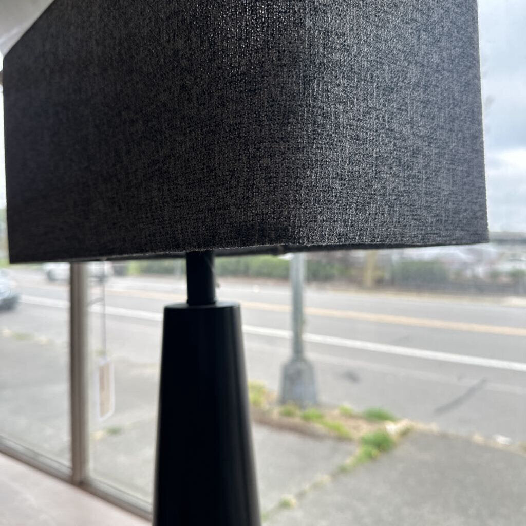Black lamp with a textured shade in front of a window with a blurred street view.